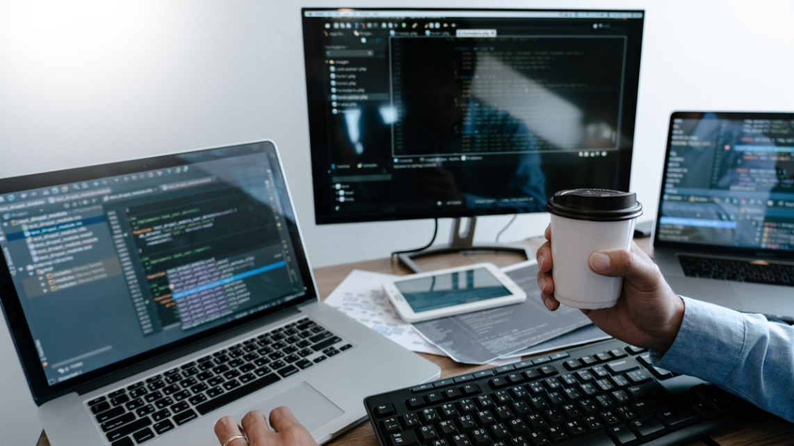 A programmer holding a cup of coffee while facing two laptops and 1 external monitor