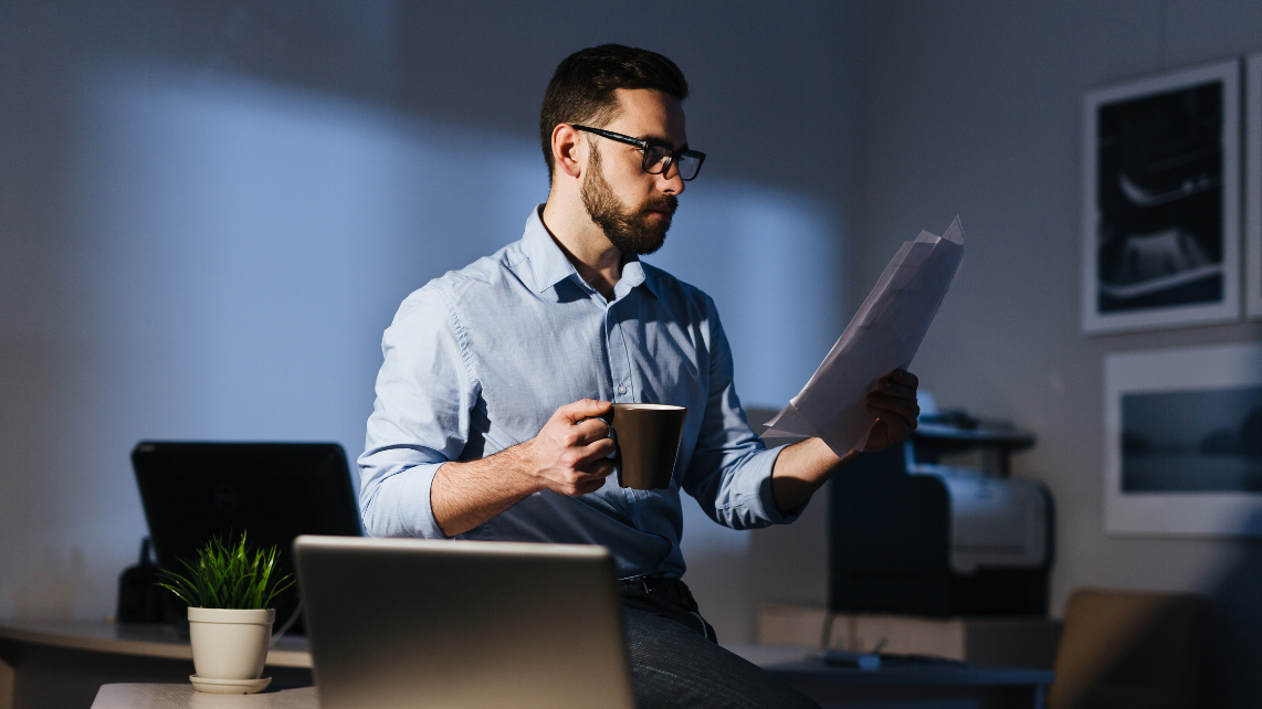 A businessman working in a dark room late at night, standing at workplace while holding papers and a cup of coffee